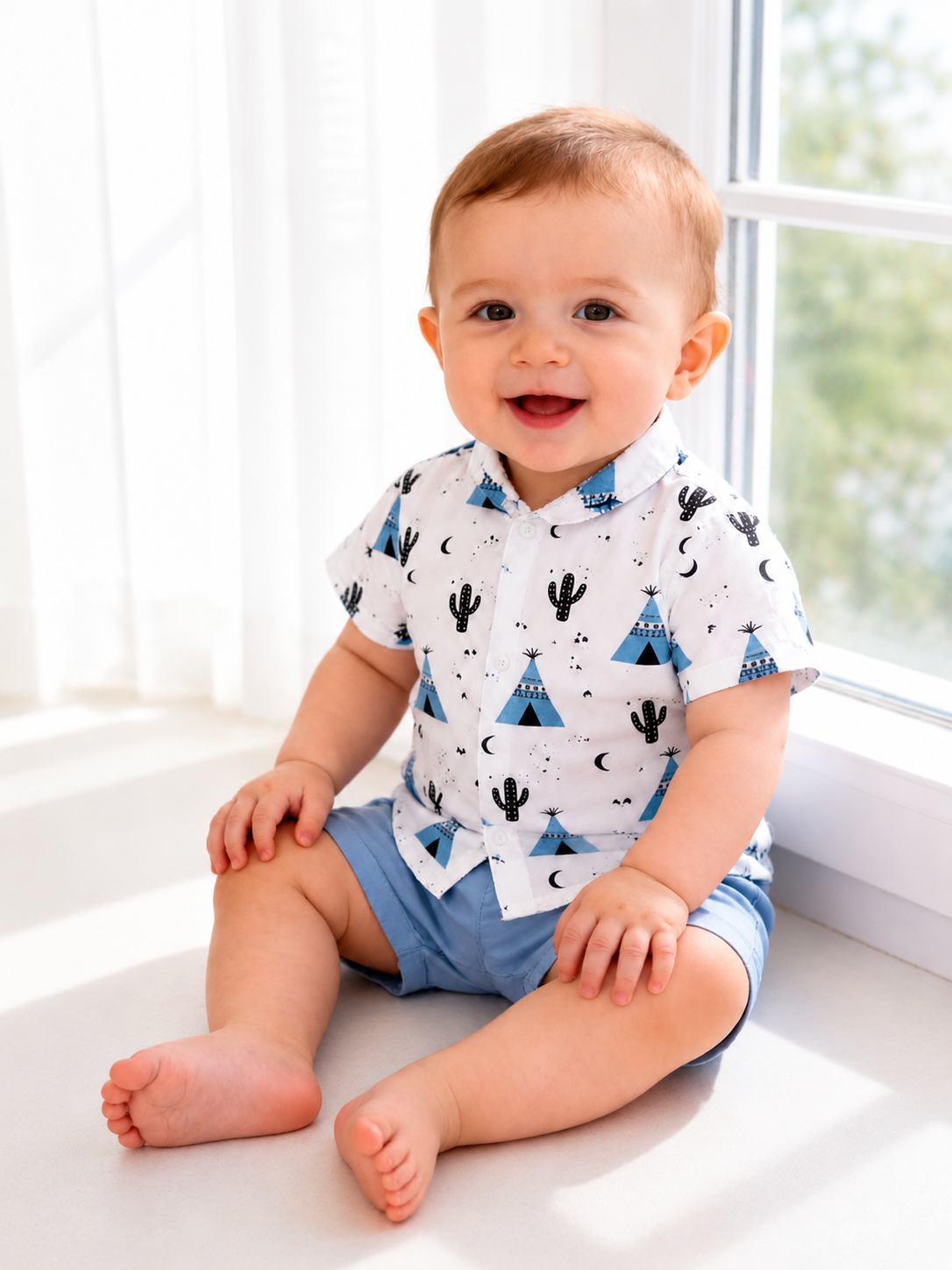 Bebê sorridente sentado próximo à janela usando conjunto infantil azul claro com camisa estampada e short confortável.